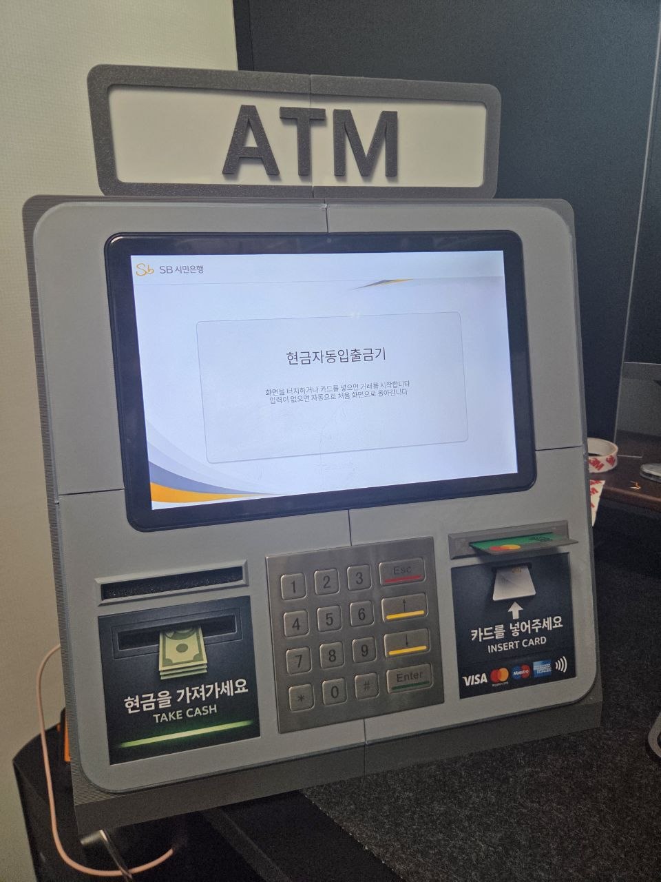 상단 ATM 표지, 터치 디스플레이, 물리 키패드, 좌측 현금 출구 모형과 우측 카드 삽입부 모형이 보이는 FlexATM 실물 시제품 전면